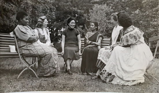 Six women chatting in a garden, dressed in their national costumes.